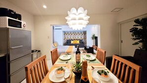 A dining area with a long table set for four, a modern chandelier, and a kitchen area with a microwave and refrigerator. - K-icon Hotels Chikusa (Nagoya)