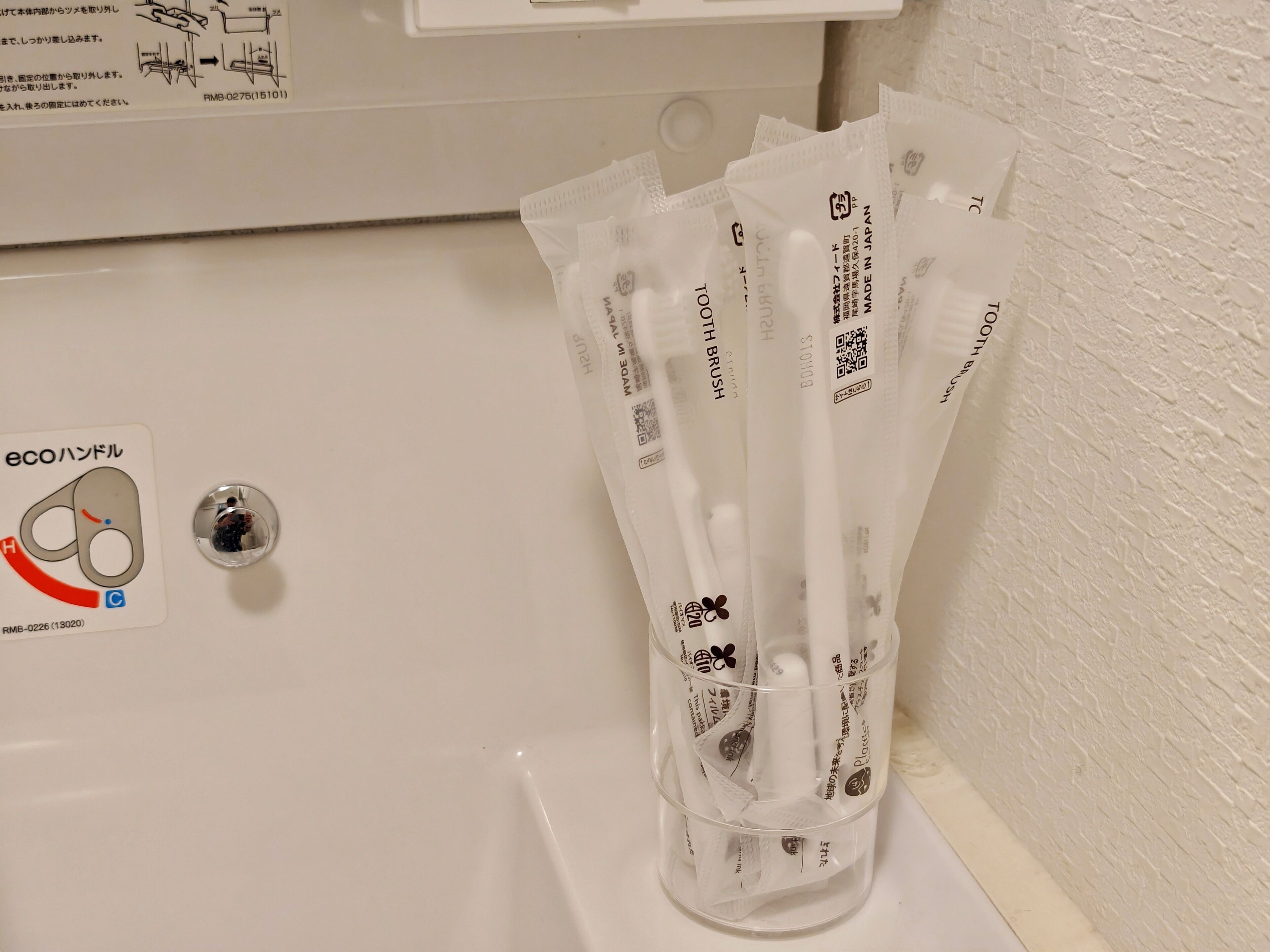 Un baño con un lavamanos blanco, dispensador de gel antibacterial y un recipiente de plástico con varios pequeños paquetes blancos, probablemente conteniendo artículos de aseo.