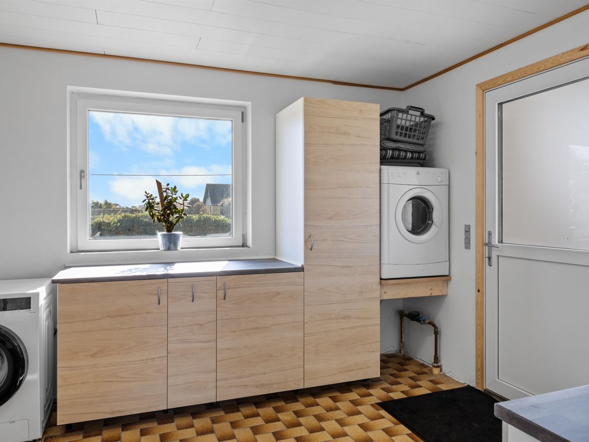 Een wasserette met een wasmachine, een droogmachine, houten kasten, een raam met uitzicht op groen, en een betegelde vloer.