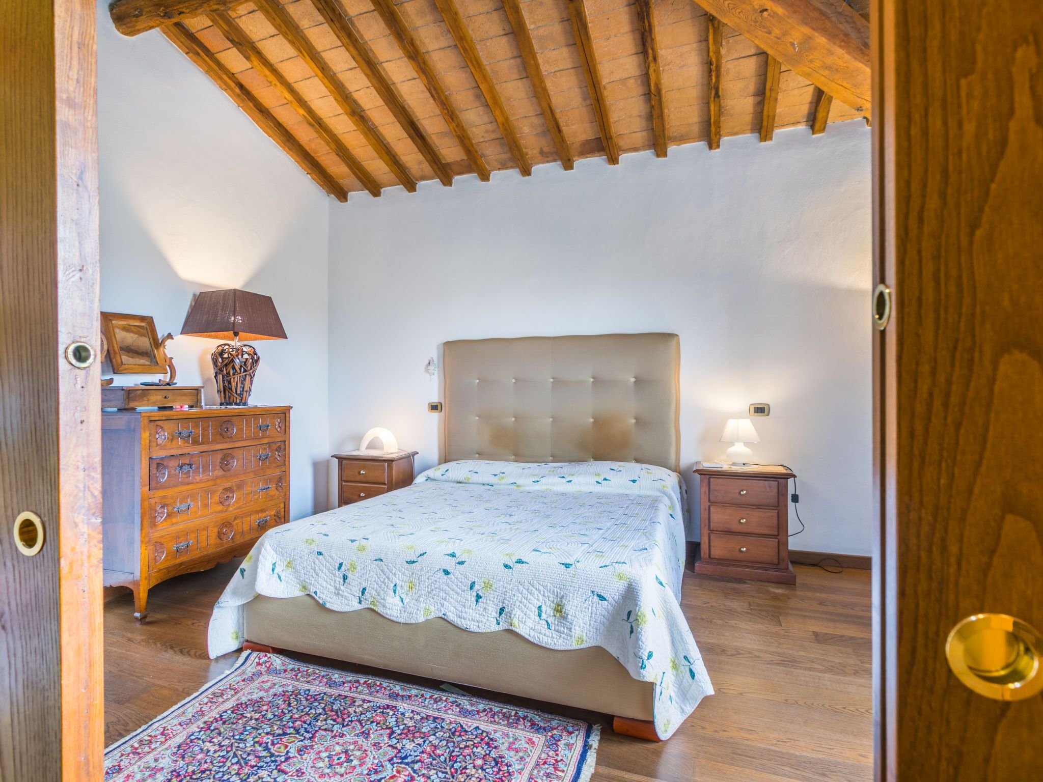 Um quarto com cama, cómoda de madeira, mesas de cabeceira e um tapete estampado.