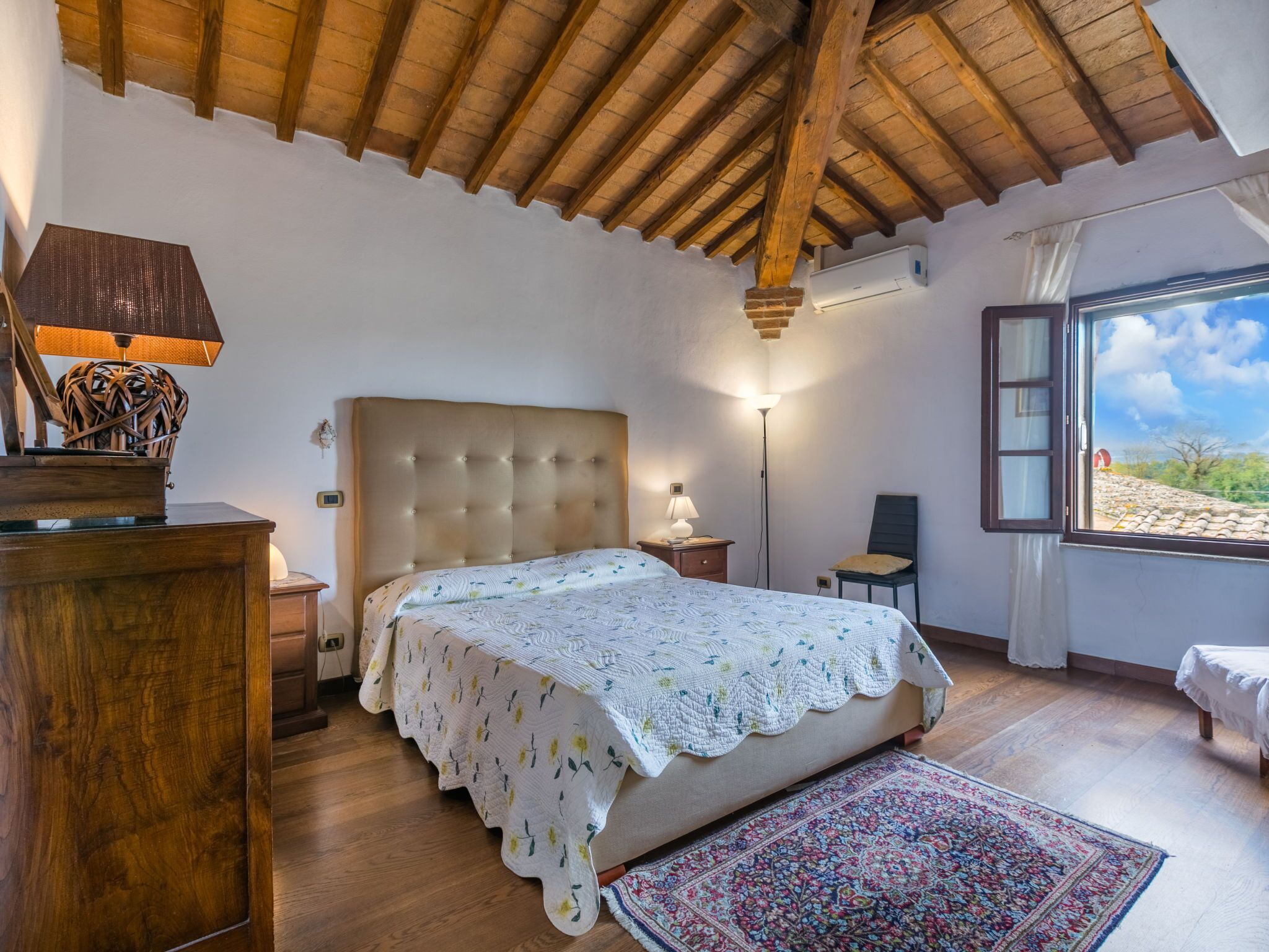 Ein Schlafzimmer mit einem großen Bett, Holzbalkendecke, einem Nachttisch, einer Lampe und Blick ins Freie.