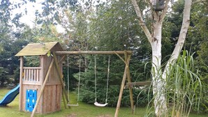 Une structure de jeux en bois, avec un toboggan et des balançoires, installée dans un espace herbeux.