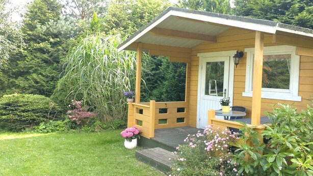 Une petite cabane en bois dotée d’un porche, entourée d’une végétation luxuriante et d’une pelouse bien entretenue.