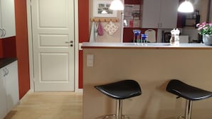 Une cuisine avec une porte blanche, deux tabourets de bar, un luminaire suspendu et un comptoir sur lequel sont disposés divers objets.
