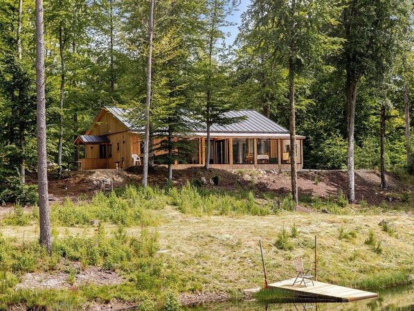 A cabin with a large deck and a covered porch, surrounded by dense forest and a small body of water. - Erikstorp 149 by Interhome (Olofström)