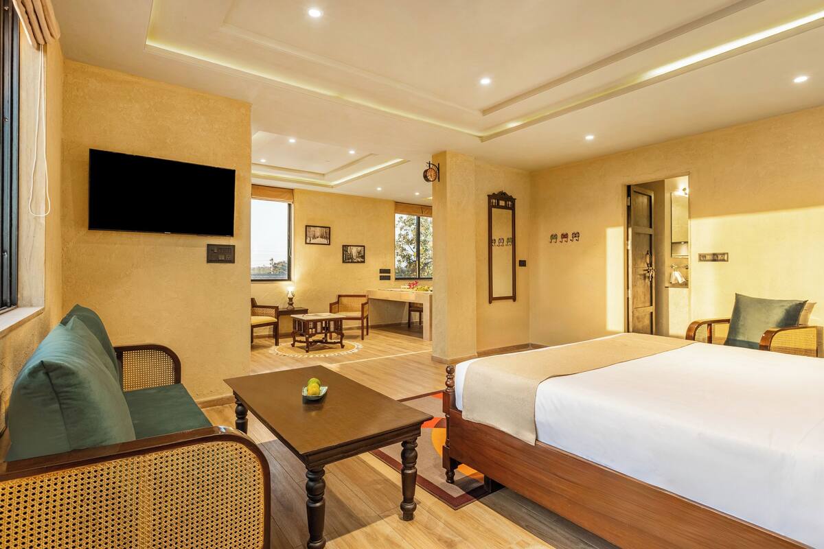 A hotel room with a large bed, a sofa, a small table with a fruit bowl, a TV mounted on the wall, and a dining area in the background.