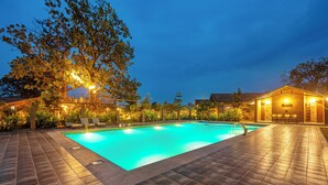 A swimming pool with a tiled deck, surrounded by trees and a building with outdoor seating. - Sterling Orient Woods Pench (Ramtek)