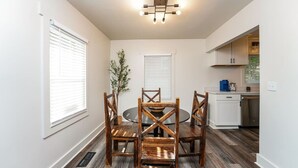 Un comedor con una mesa redonda y cuatro sillas de madera, una planta pequeña y una cocina con muebles blancos y lavavajillas.