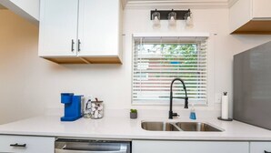 Una cocina moderna con armarios blancos, un lavamanos de acero inoxidable, lavavajillas y una ventana con persianas.