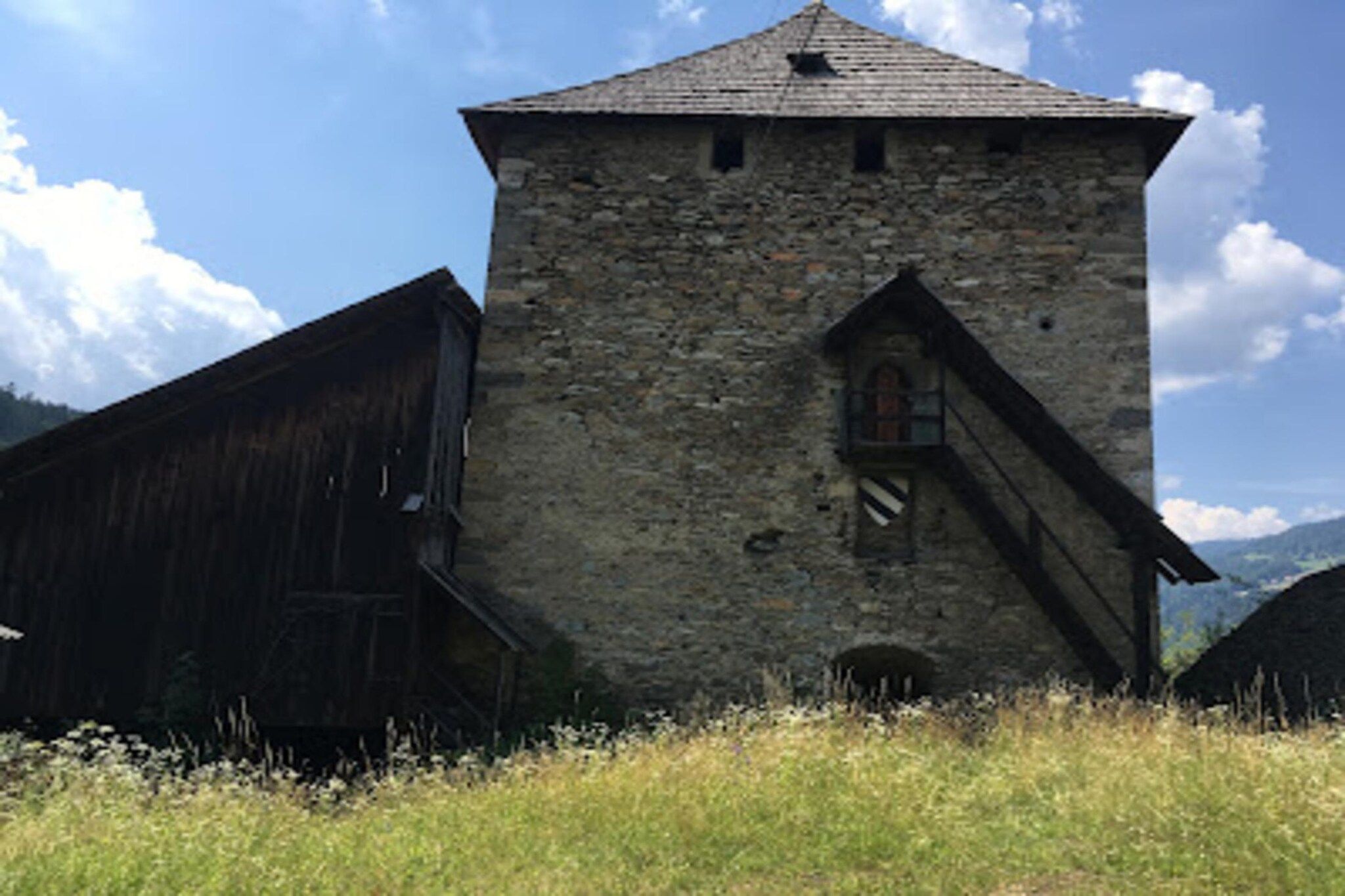 A stone tower with a steeply pitched roof, a wooden staircase leading to a door, and a rustic wooden building to the left.