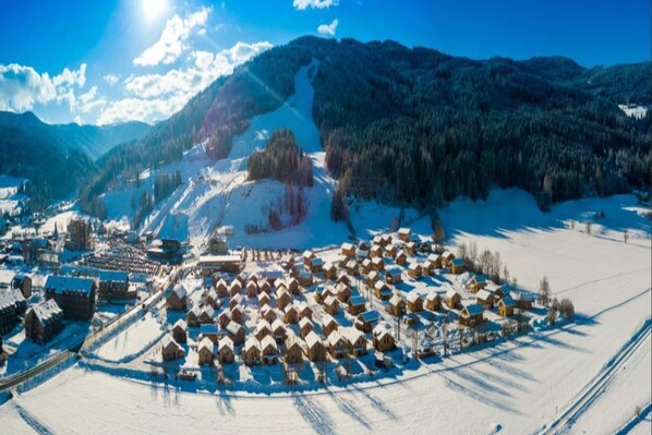 Un pueblo cubierto de nieve con casas de madera tradicionales, una iglesia y una pista de esquí al fondo.