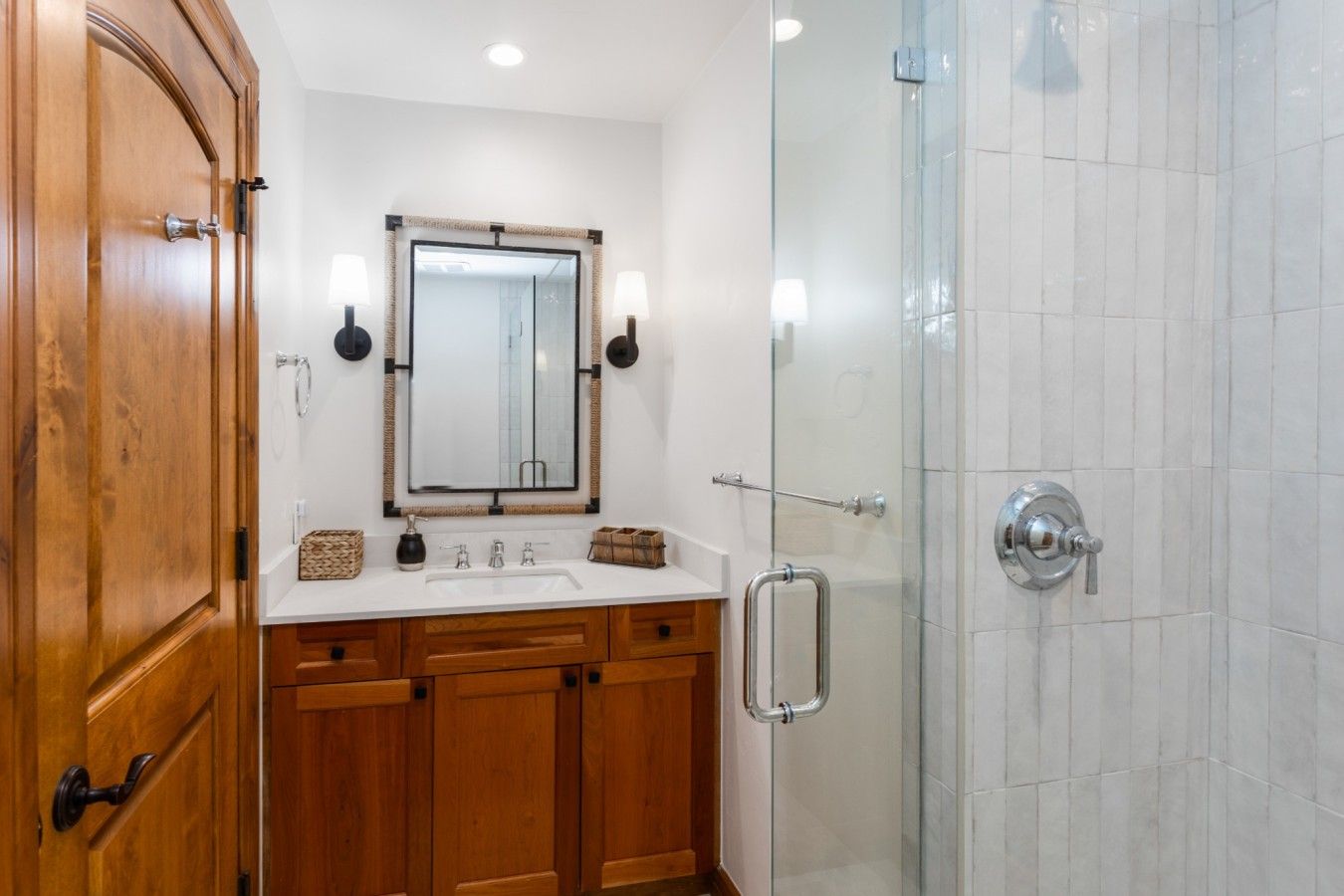 Un baño con armarios de madera, un lavamanos blanco, un espejo con apliques de pared y una cabina de ducha de vidrio.