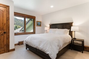 A bedroom with a large bed, two bedside tables with lamps, a window with a view of a mountain, and a wooden door.