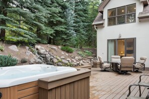 Un bain à remous sur une terrasse en bois, agrémentée de mobilier d’extérieur, entouré d’un jardin aménagé et d’une maison dotée d’une grande fenêtre.