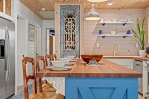 A kitchen with a wooden island, chairs, a refrigerator, a sink, and shelves with dishes.