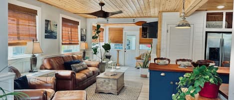 A spacious living room with a leather sofa, a coffee table, and a kitchen area with a blue cabinet and a hanging light fixture.