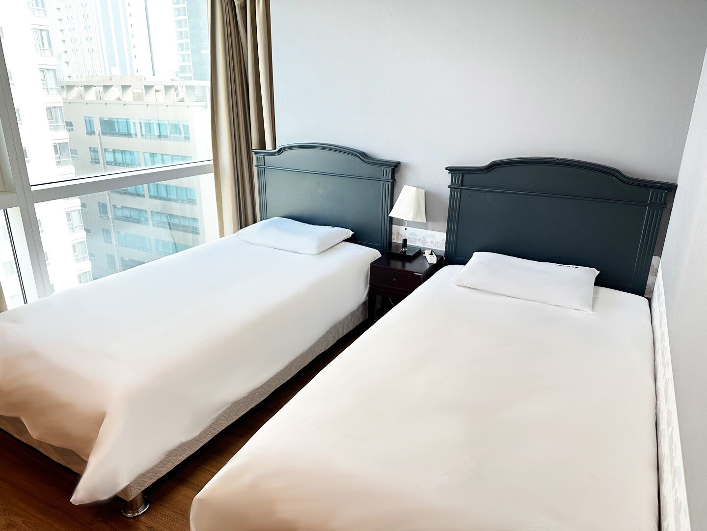 Ein Hotelzimmer mit zwei Einzelbetten, einem Nachttisch mit Lampe und einem Fenster mit Blick auf Gebäude.