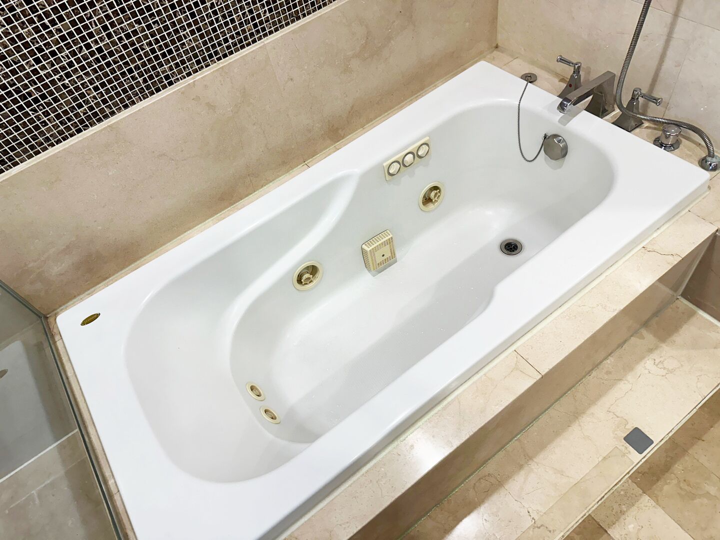 Una bañera blanca con múltiples grifos y un cabezal de ducha manual, frente a una pared de azulejos con un diseño tipo cuadrícula.