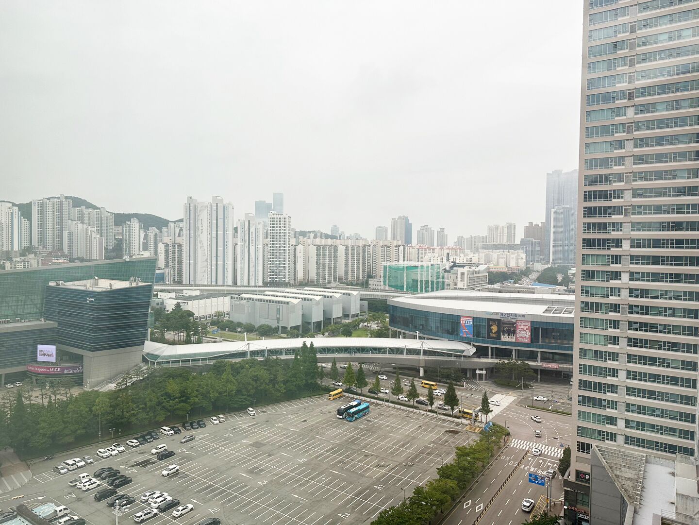 A parking lot with multiple cars and a bus, surrounded by modern buildings and a high-rise structure.