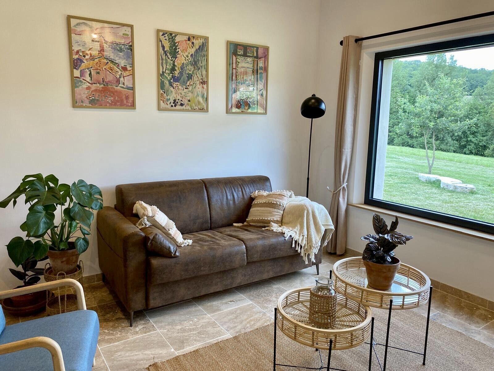 Un salon avec un canapé en cuir marron, un fauteuil bleu et une grande fenêtre donnant sur un paysage verdoyant. Trois tableaux ornent le mur, et une grande lampe sur pied se dresse dans un coin.
