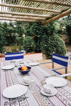 Ein Essbereich im Freien mit einem für vier Personen gedeckten Tisch, umgeben von Stühlen und einer überdachten Pergola aus Bambus.