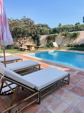 Ein Poolbereich mit Liegestühlen und einem Tisch, umgeben von viel Grün und blauem Himmel.