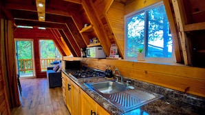 A wooden cabin interior with a kitchen, dining area, and a view of the outdoors.