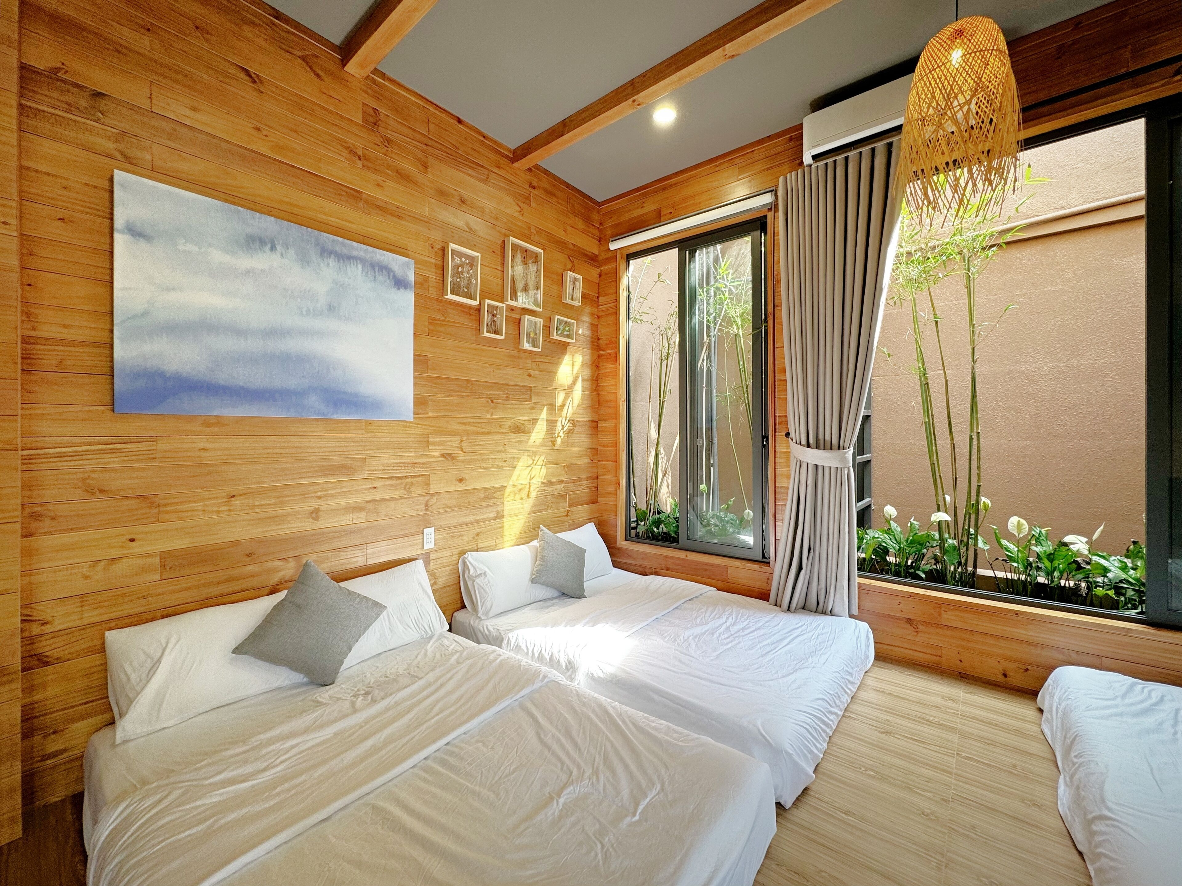 A room with wooden walls, two beds with white linens, a large window with a view of greenery, and a hanging light fixture.