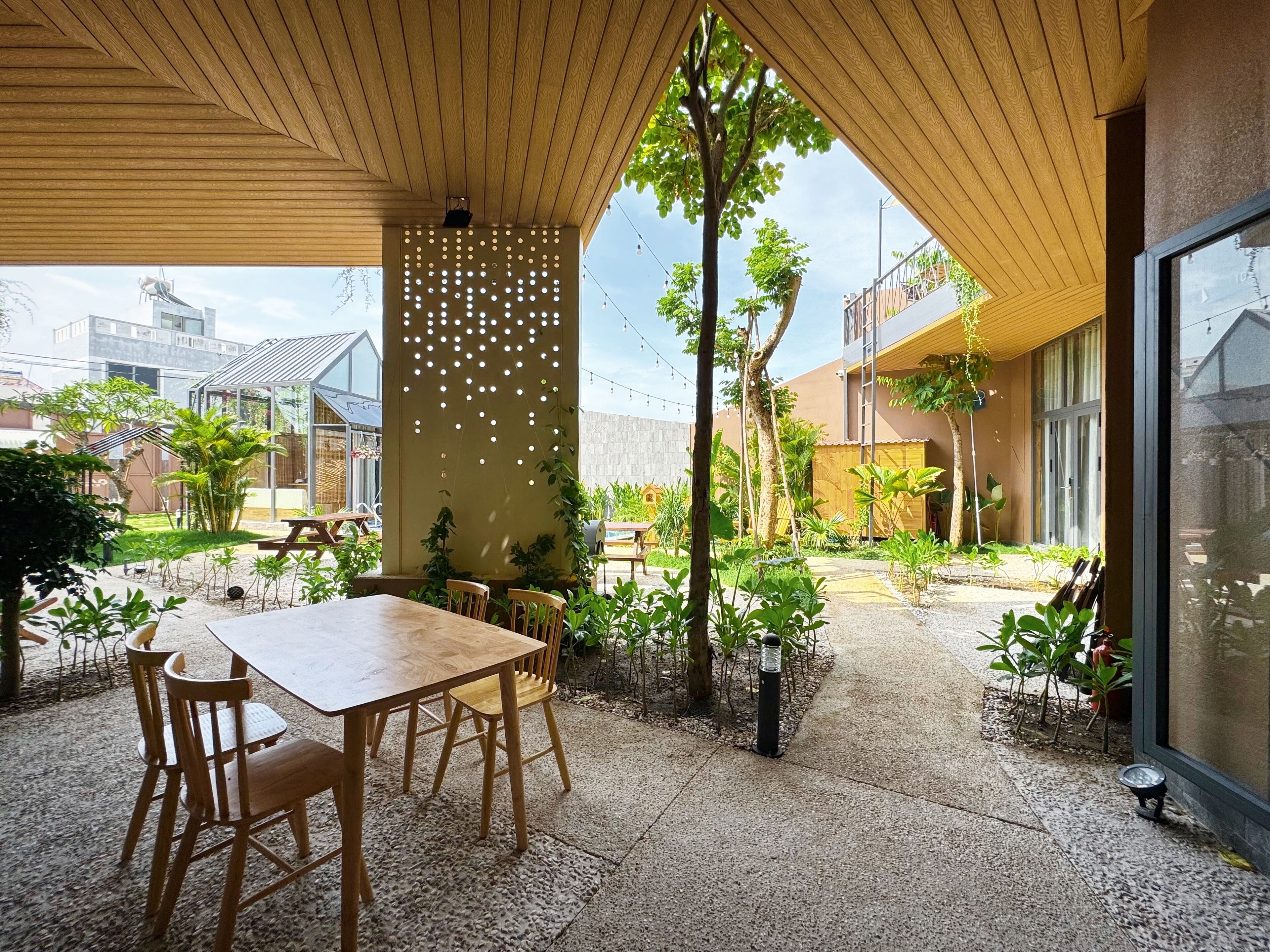 Un patio al aire libre con una mesa y sillas de madera, rodeado de macetas con plantas y una pared decorativa con luces.