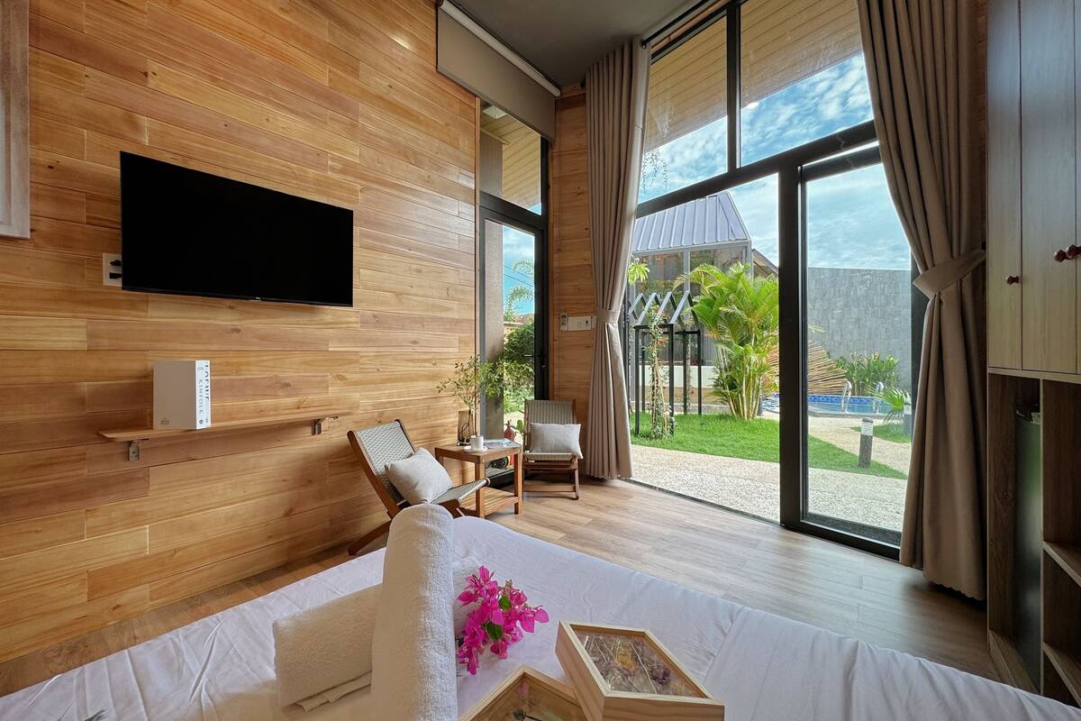 A modern hotel room with a wooden wall, a flat-screen TV, a bed with white linens, a small table with a cup, and a view of the outdoors.