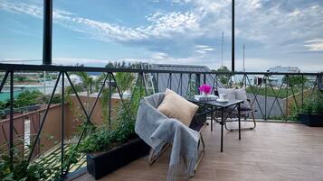 A balcony with a view of the city, a table with a book and a cup, and a chair with a blanket.