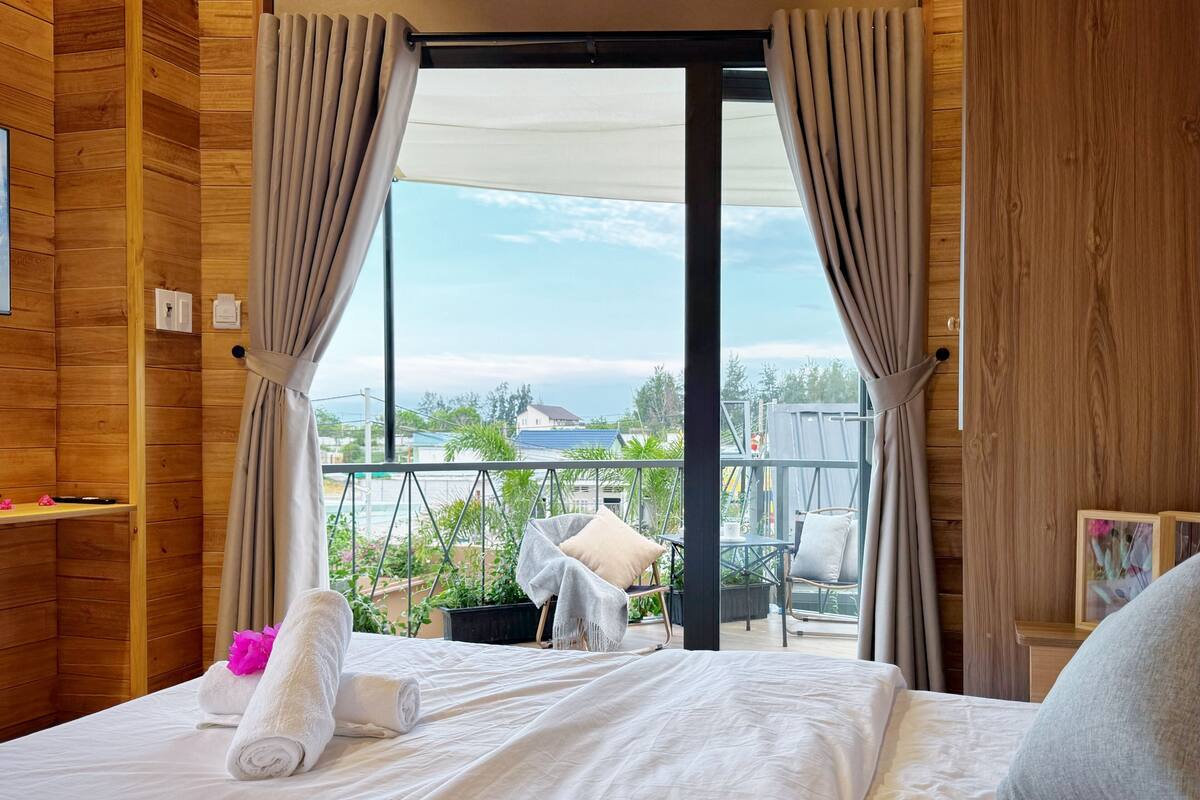 A hotel room with a balcony, wooden walls, a bed with white linens, and a view of the outside.