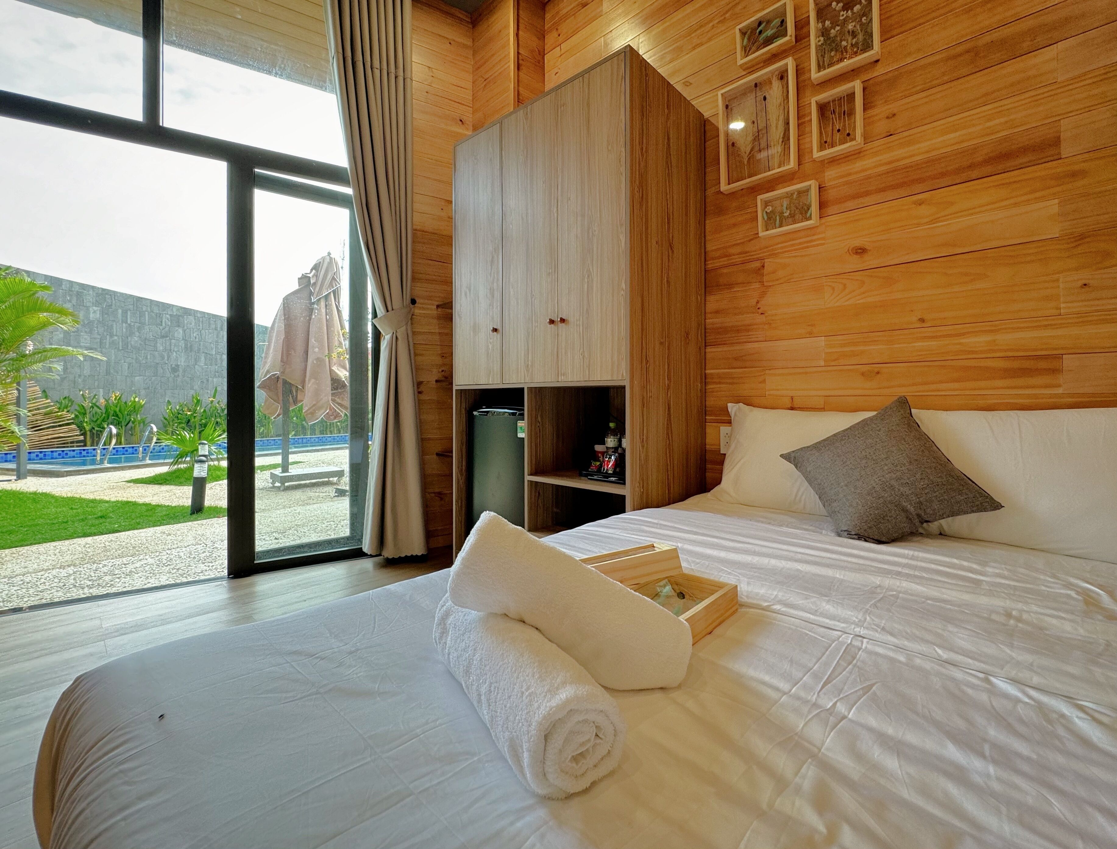 A hotel room with a large bed, a wooden wardrobe, a small shelf, and a view of a pool and garden.
