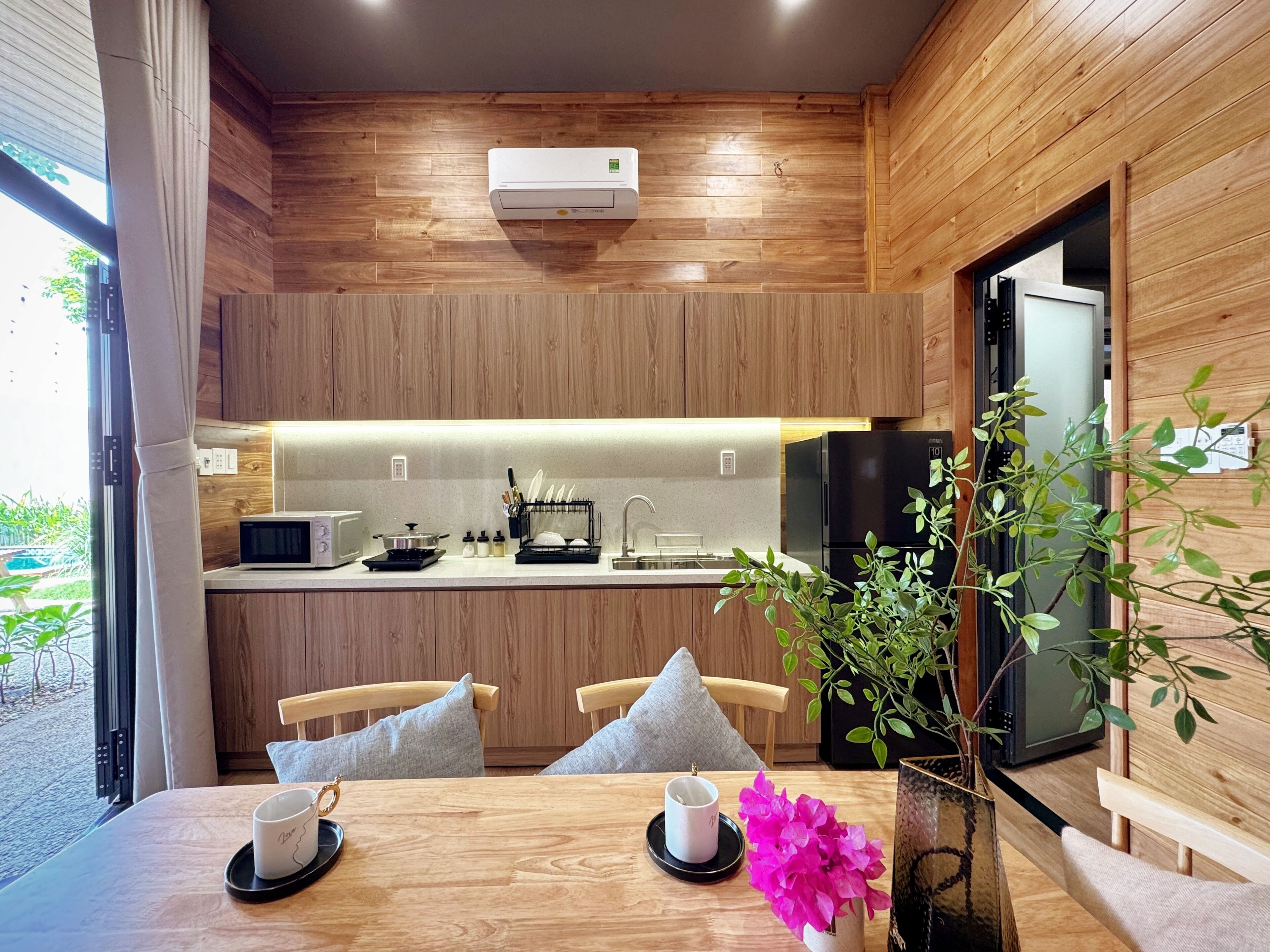 Una cocina moderna con armarios de madera, un microondas integrado, una estufa y un refrigerador. Hay un comedor con una mesa, sillas y un florero con flores.