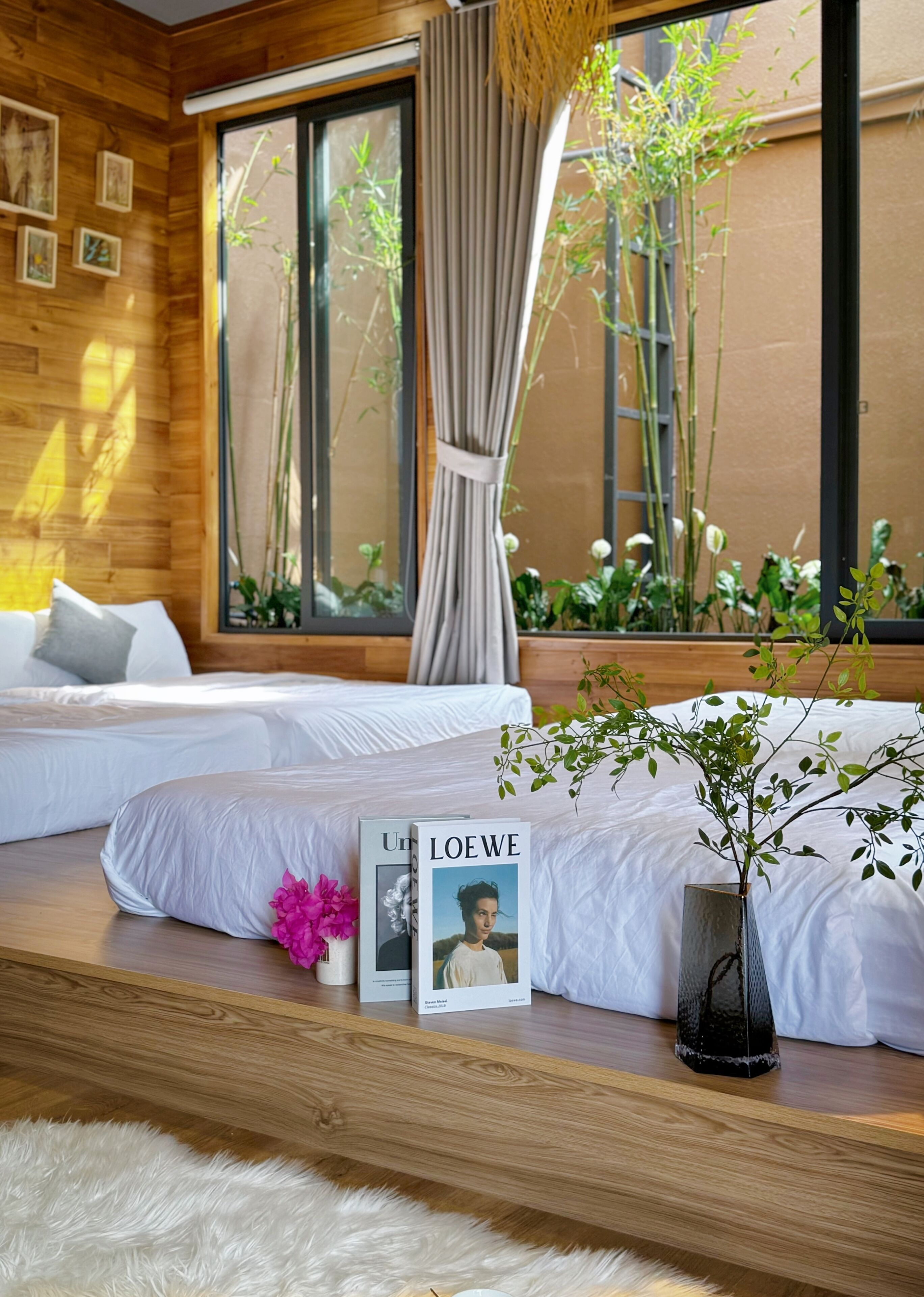 A modern bedroom with two beds, a large window with bamboo plants, and a bookshelf with books and a vase.