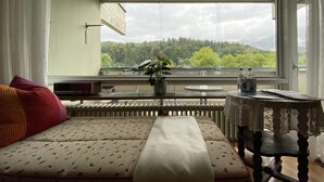 Une pièce comprenant un canapé, une petite table, un radiateur et une fenêtre donnant sur des arbres.