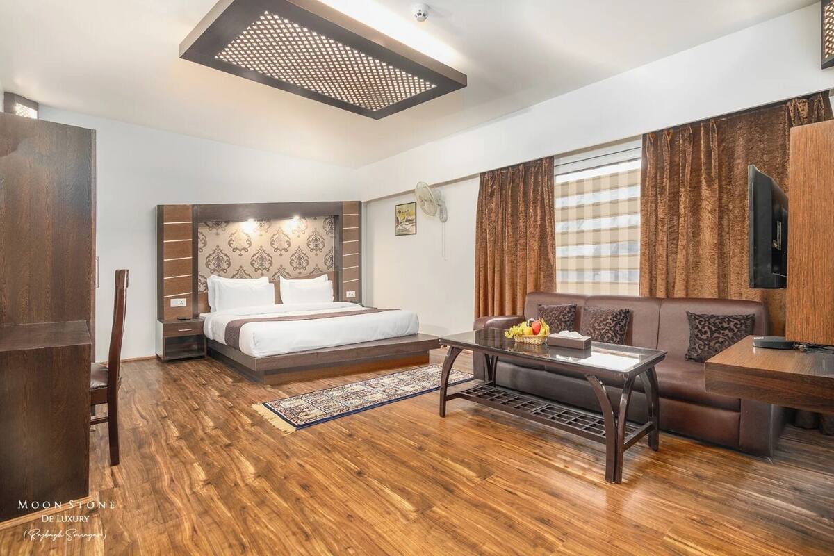 A modern hotel room with a wooden floor, a bed with a headboard, a sofa, a coffee table, and a TV mounted on the wall.