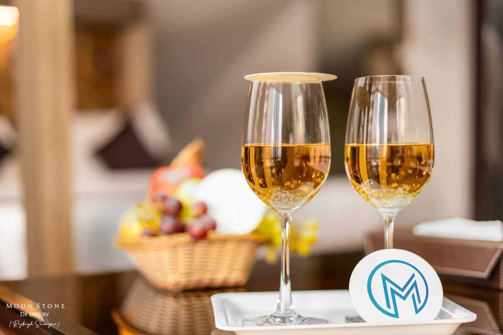 Two glasses of white wine on a tray with a logo, a basket of fruit, and a wicker table.