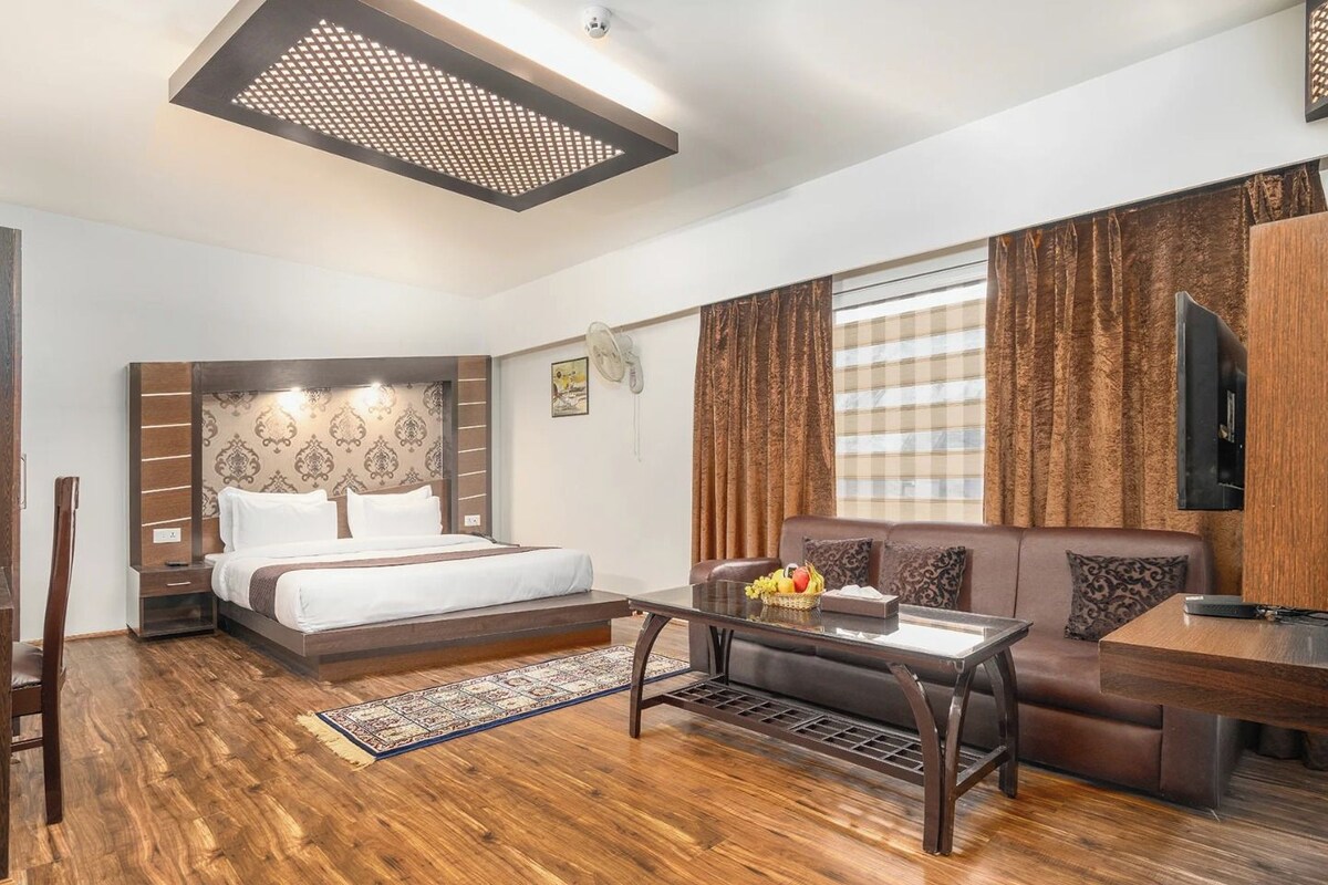 A modern hotel room with a wooden floor, a bed with a headboard, a sofa, a coffee table, and a TV mounted on the wall.