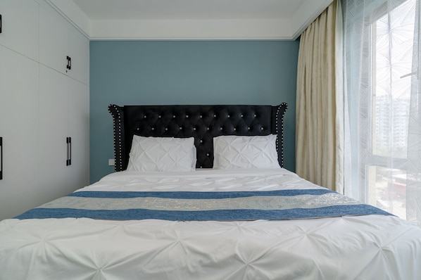 A bedroom with a tufted headboard, white bedding, and a view of the city through sheer curtains. - ALBA Garden 1BR with Gym & Infinity Pool (Nairobi)