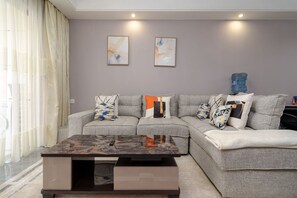 A modern living room with a grey sectional sofa, a glass coffee table, and two abstract paintings on the wall. - ALBA Garden 1BR with Gym & Infinity Pool (Nairobi)