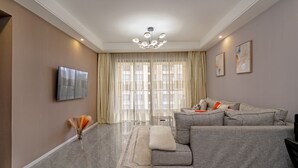 Een moderne woonkamer met een grijze modulaire bank, een flatscreen televisie, een kroonluchter en een groot raam met dunne gordijnen.