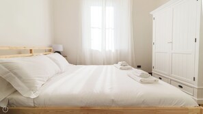 Un letto rifatto con lenzuola e cuscini bianchi, una lampada da comodino, una finestra con tende leggere e un armadio bianco.