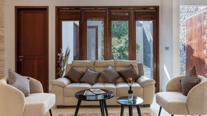 Ein modernes Wohnzimmer mit einem beigen Sofa, zwei Sesseln, einem Couchtisch und einem großen Fenster mit Holzhörchen.