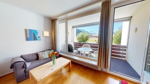 A modern living room with a sofa, a wooden coffee table, and a view of a balcony with outdoor furniture.