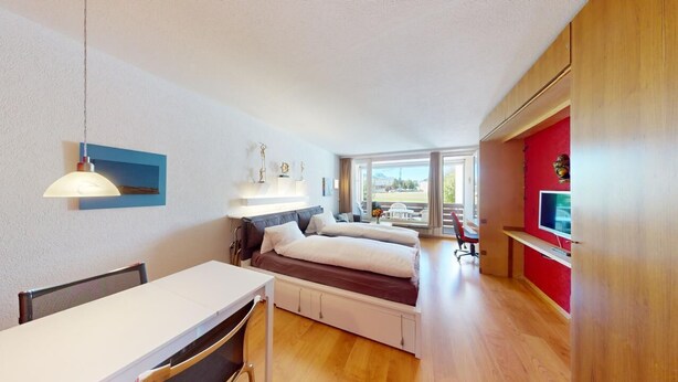 A compact room with a bed, a desk, and a chair. There is a window providing natural light and a view of the outdoors.