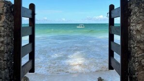 Una entrada a la playa con un portón de madera, agua turquesa cristalina y un barco a lo lejos.