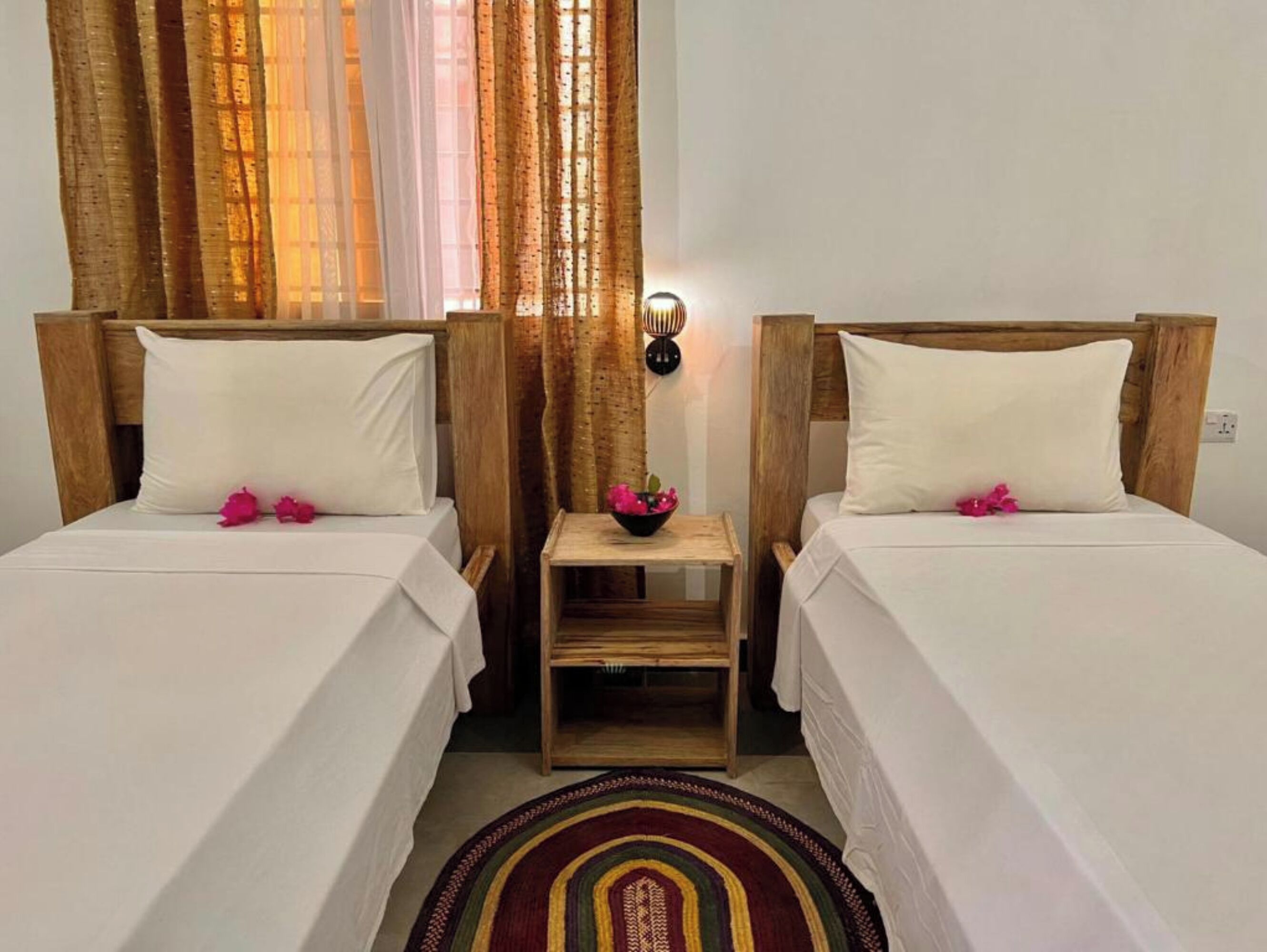 A room with two beds, each with white linens and pink flowers. There is a small wooden nightstand between the beds with a bowl of flowers. The room has wooden headboards and a light fixture on the wall.