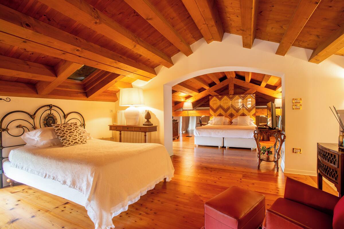 Ein geräumiges Schlafzimmer mit Holzbalkendecke, einem großen Bett, einer Sitzecke mit einem roten Sessel und Blick in einen weiteren Raum.