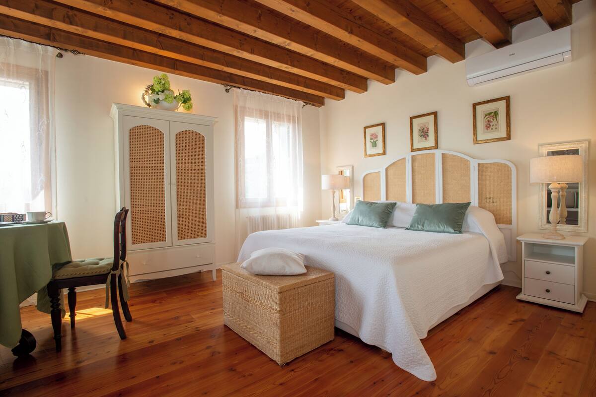 Ein Schlafzimmer mit einem großen Bett, einem Korbstuhl, einem kleinen Tisch mit grüner Tischdecke und einem weißen Schrank.